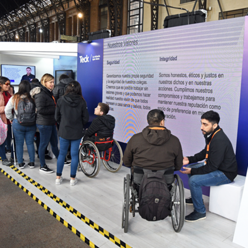 People at an event near a screen displaying values; some individuals are in wheelchairs.